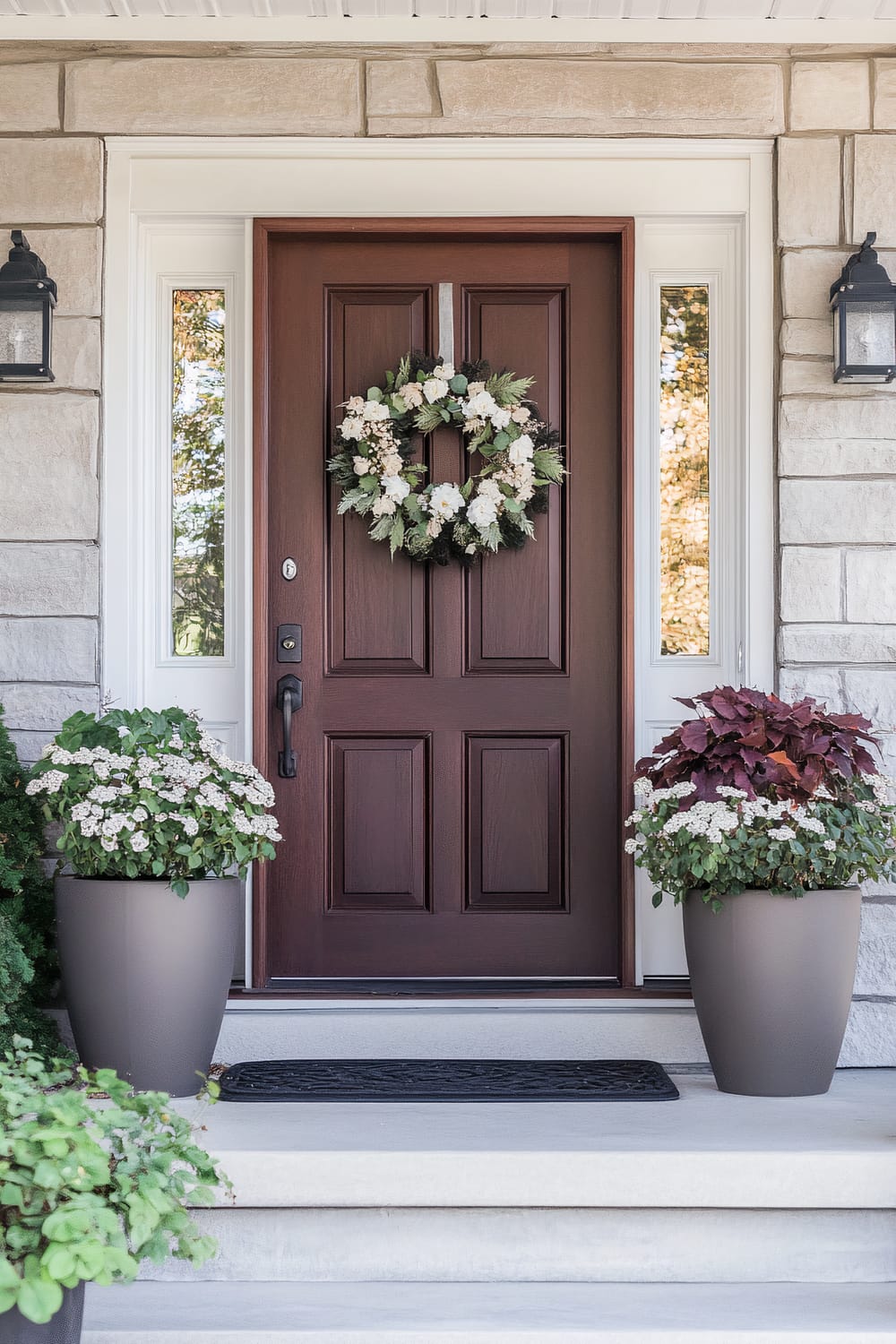 A front door designed with a dark wooden panel adorned with a lush wreath made of white flowers and green foliage. The entrance is flanked by two small windows with white trim and beige stone walls. Black wall-mounted lanterns are placed symmetrically on either side. Two large gray planters with green and maroon foliage plants are placed on either side of the door, atop three steps leading to the entrance.