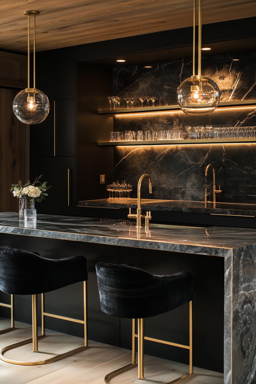 Modern kitchen featuring stone countertops, brass fixtures, and glass globe pendant lights. The countertops are a dark, marbled stone with a polished finish. Open shelving with a backlit black marbled backsplash displays glassware. Two velvet-upholstered bar stools with gold frames are placed at the island.