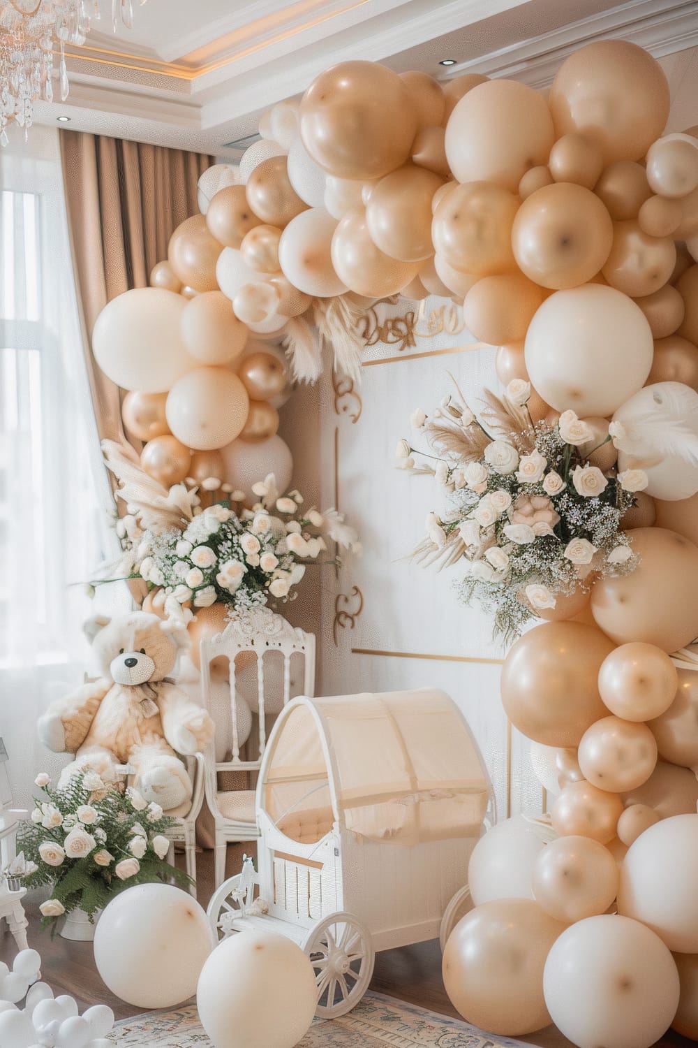 The image shows an elegantly decorated room for a baby's event, with a neutral color palette dominated by beige and white. The scene features numerous inflated balloons in shades of cream and gold forming an arch around a decorative white door. There are also lush floral arrangements consisting of white roses, baby’s breath, and pampas grass. A large, plush teddy bear, a vintage-style white chair, and a small white baby carriage with a canopy are prominently positioned in the room. A luxurious chandelier with hanging crystals can be seen on the ceiling, illuminated by soft, natural light filtering through the window with beige curtains.