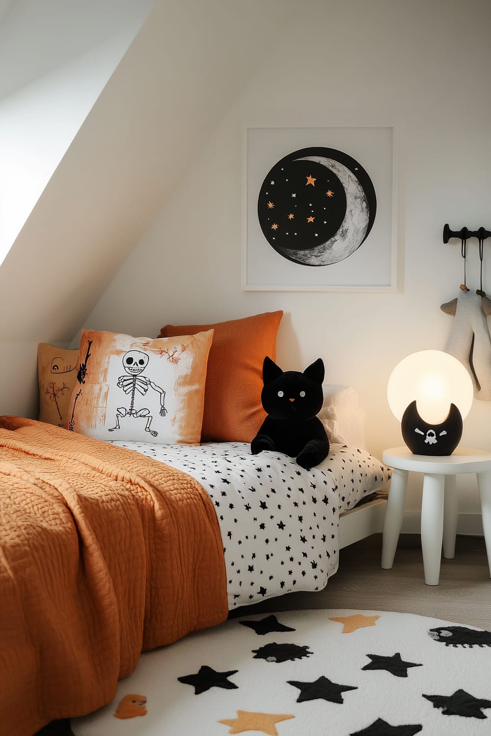 A child's bedroom with a Halloween theme. The bed has an orange blanket and white bedsheets with black stars. There are several pillows, one decorated with a skeleton and another in solid orange. A black cat plush toy sits on the bed. A round nightstand next to the bed holds a lamp shaped like a black crescent moon with a skull and crossbones, illuminating a globe light. A framed picture of a crescent moon and stars hangs on the wall above the bed. A white rug with black and orange stars is on the floor.