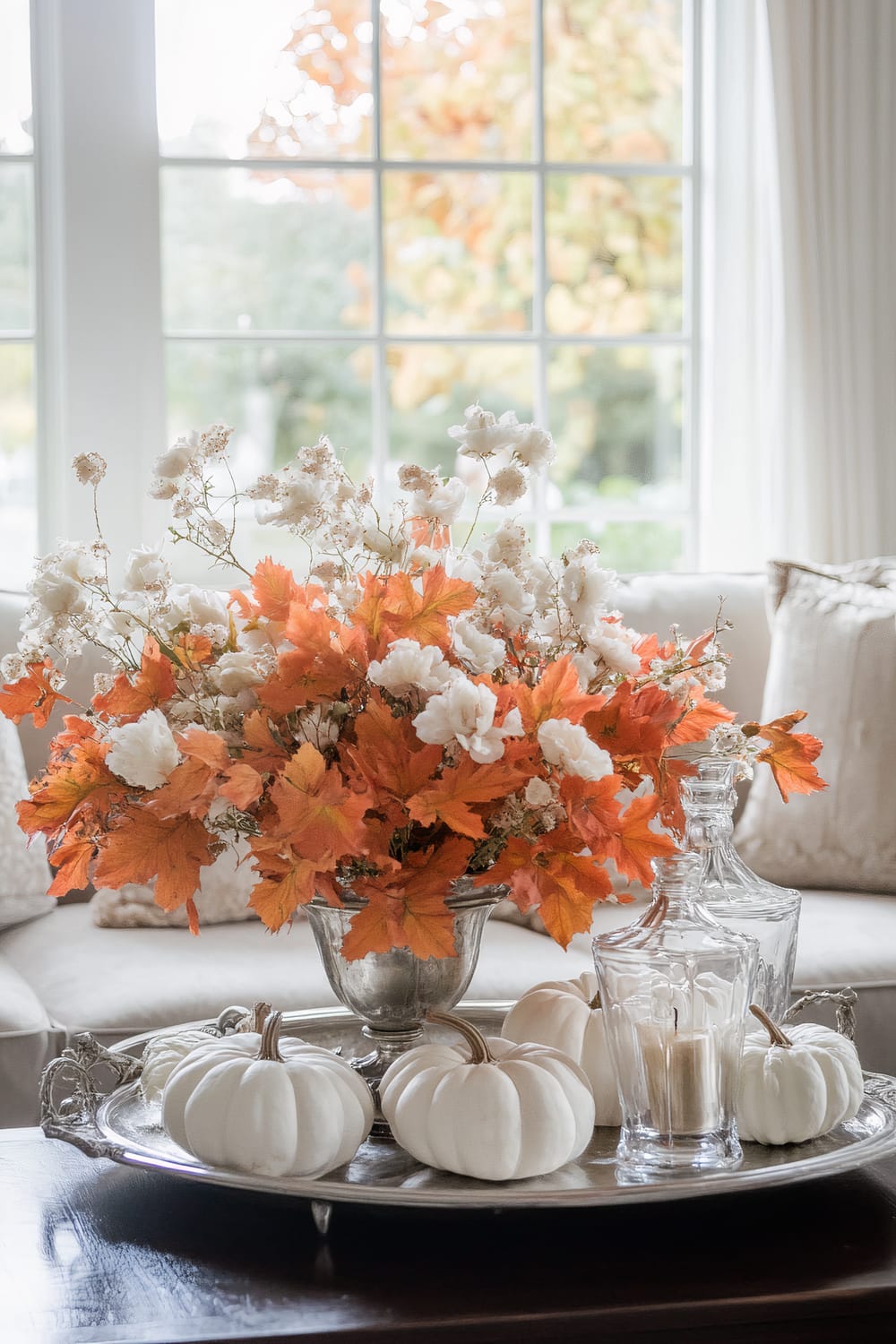 An elegant arrangement of white and orange autumn leaves with white flowers in a decorative vase is placed centrally on a living room coffee table. Surrounding the vase are mini white pumpkins and glass decanters, sitting on an antique silver tray. Large windows in the background offer a view of colorful fall foliage.