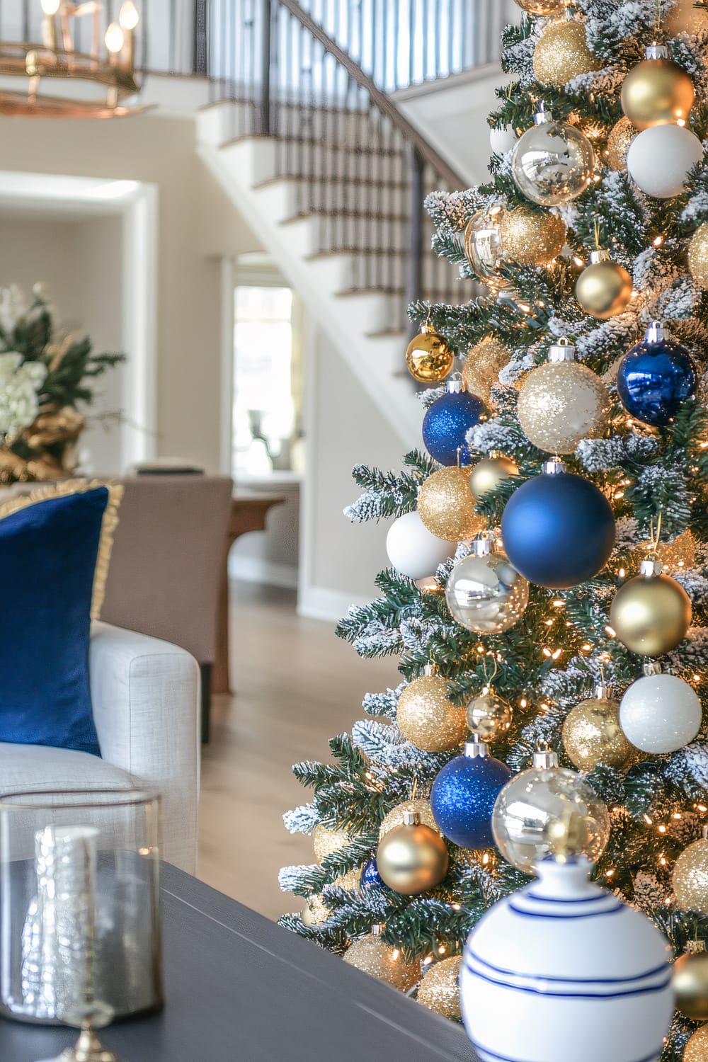 A beautifully decorated Christmas tree stands in a sophisticated living room. The tree is adorned with glittering blue, gold, white, and silver ornaments, some of which are reflective, surrounded by twinkling fairy lights. The branches have a snow-frosted appearance. In the foreground, a dark wooden coffee table holds a glass candle holder and a white decorative container with blue stripes. Behind the tree, a staircase with dark wooden railings curves upwards, with a soft light filtering in through a window in the background. The interior is elegantly staged with neutral tones and plush furnishings, including a gray sofa with a blue cushion.