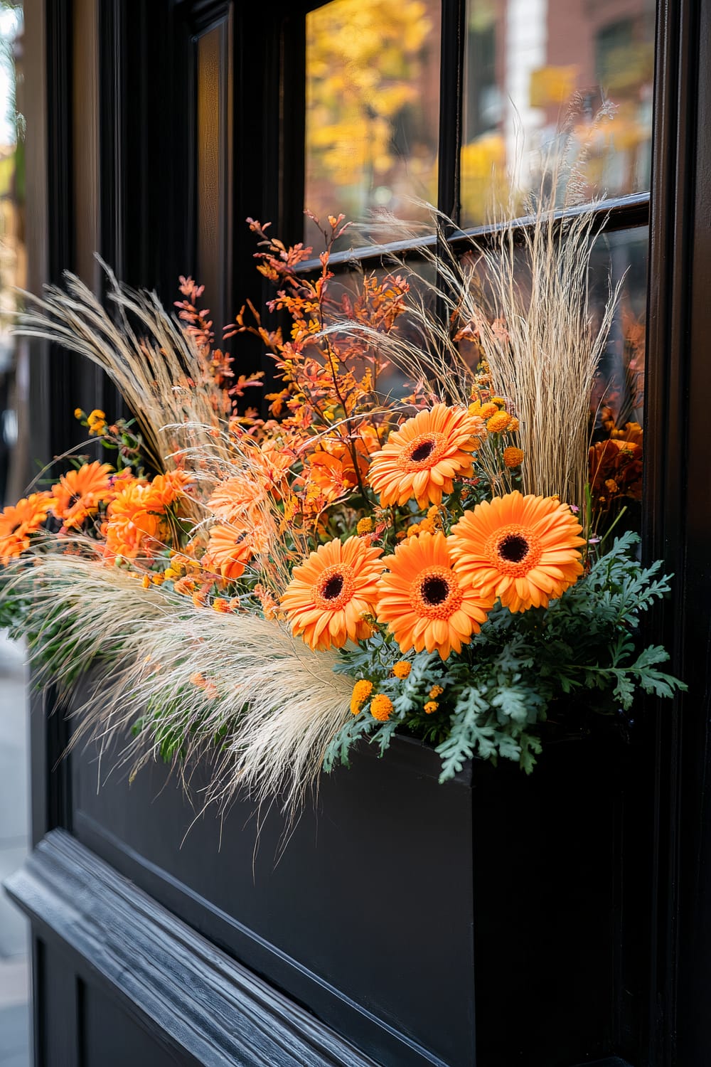 A vibrant flower box filled with striking orange gerbera daisies and leafy green foliage is mounted on a black window sill. The display is complemented by feathery ornamental grasses and delicate orange blooms, enhancing the autumnal scene. The background reflects an urban street blurred with the warm amber hues of fallen leaves.