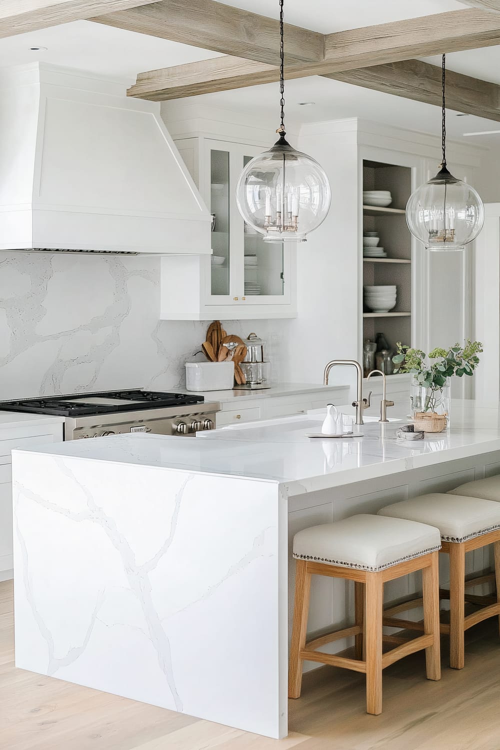 A modern design kitchen featuring a large white island with a marble pattern and bar stools with light wooden frames and upholstered seats. The island includes a sleek built-in steel sink and a contemporary faucet. Above it hang two clear glass pendant lights with ornate candle-style bulbs. Behind the island, the counter extends with a gas stove, a backsplash with matching white marble patterns, a hood, and built-in shelving with glass-front cabinets displaying crockery and kitchenware. Decorative beams in a light wood finish adorn the ceiling, adding a rustic touch.