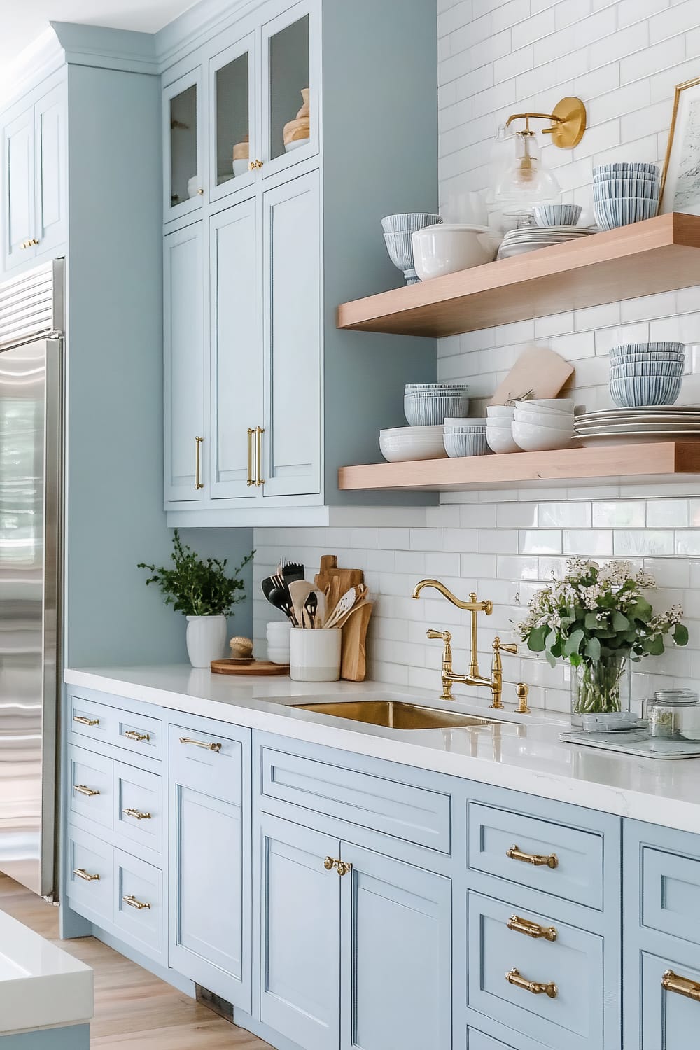 This image features a sophisticated kitchen interior with pastel blue cabinets and drawers adorned with elegant gold handles and fixtures. The back wall consists of clean, white subway tiles. Two wooden open shelves display a variety of white and grey ceramic bowls and plates. A brass faucet elegantly complements the cohesive design, and a lush bouquet of white flowers in a glass vase adds a touch of nature. The countertops are white with subtle grey veining, enhancing the overall airy and refined ambiance.