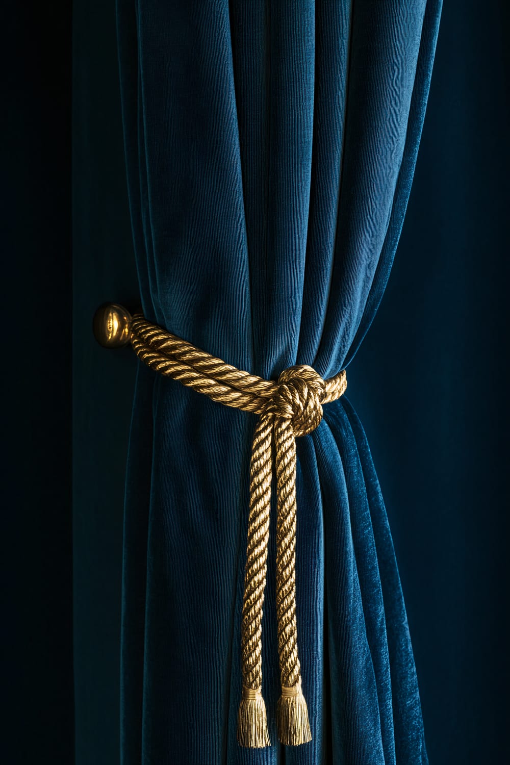 A close-up view of a luxurious, deep blue velvet curtain tied back with an elegant, thick, golden rope. The rope is knotted and features tassels at the end, adding to the opulent appearance.