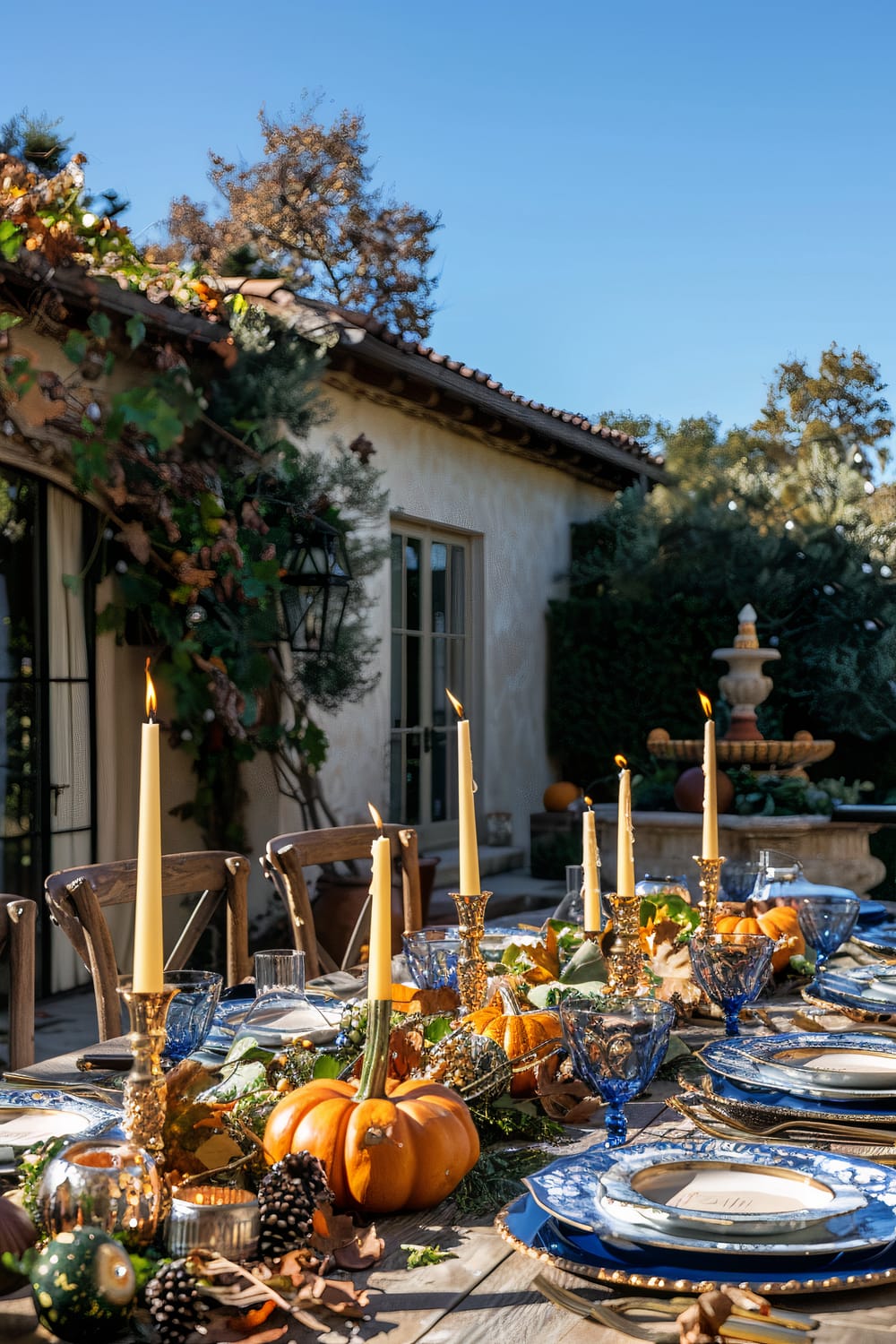 A beautifully set outdoor dining table adorned with autumn-themed decorations. The centerpiece features small orange pumpkins, pine cones, and leaves, along with flickering tall candles in ornate holders. Elegant blue and white china plates, gold-rimmed glasses, and crystal glassware complement the rustic wooden table. The background showcases a stucco building with hanging ivy, a classical fountain, and vibrant greenery, all bathed in warm, natural sunlight.