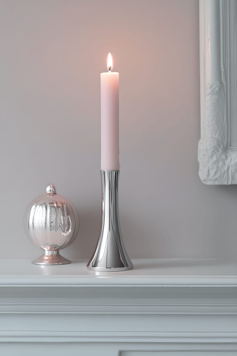 A modern silver metal candle tower with a lit pastel-pink candle positioned on a white minimalist mantel. The warm flame casts a soft pink light on the structure, transforming it into a romantic centerpiece.