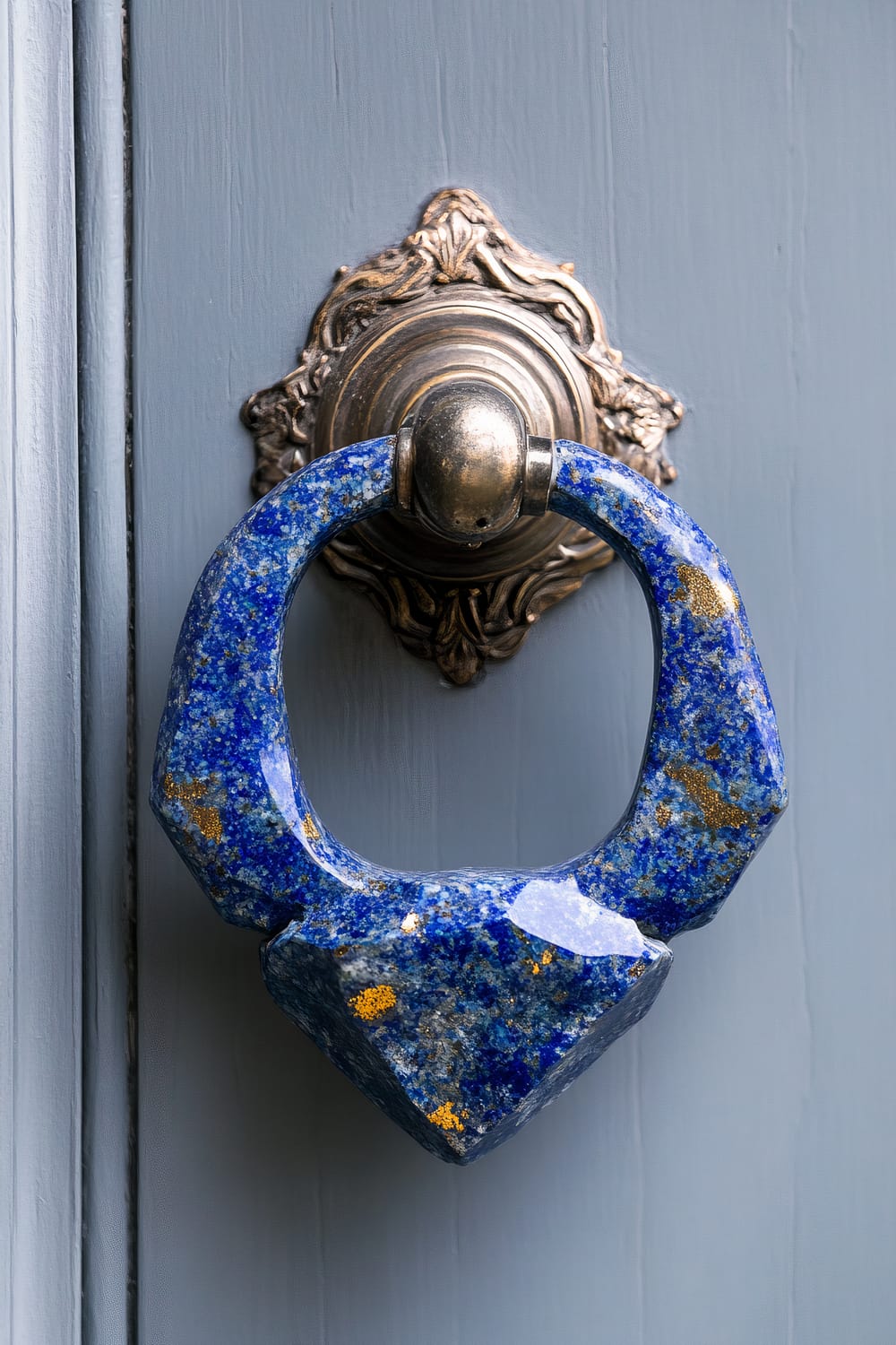 A close-up view of a blue stone door knocker with gold flecks, mounted on a weathered, ornate brass backplate attached to a light blue painted wooden door.