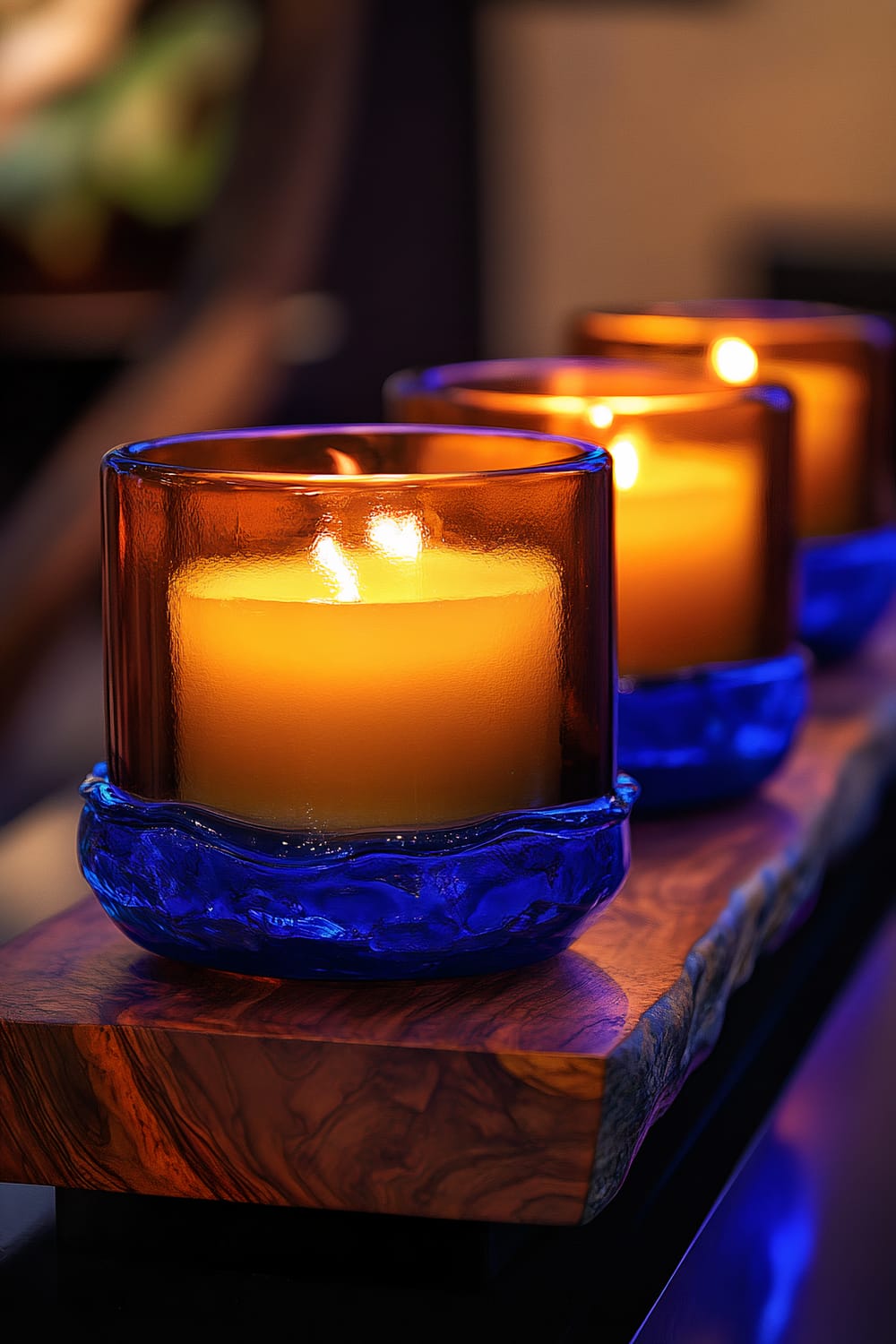 Three amber-colored candles are arranged in a row within cobalt blue glass holders. These are set on a dark wooden base with prominent grain patterns. The candles are lit, emitting a warm glow that contrasts beautifully with the blue glass and the dark wood. The melted wax of the candles adds texture to the scene.