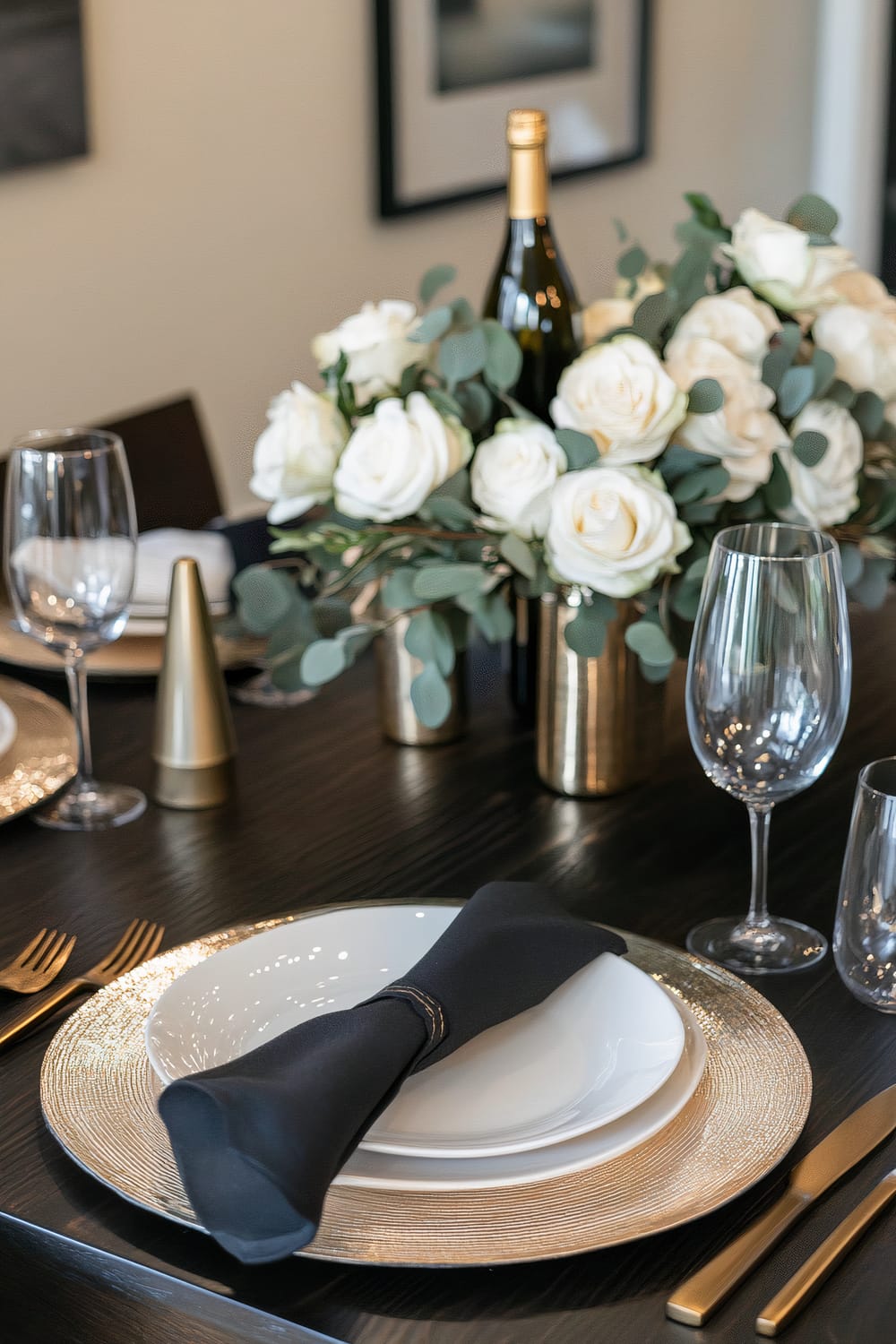 An elegantly set dining table features a central arrangement of white roses and eucalyptus in golden vases. Around the table, there are white plates on golden chargers, paired with black napkins and sleek golden cutlery. A bottle of wine is visible among the floral arrangement, and the table also includes wine glasses and gold decorative cone-shaped items, set against a dark wood tabletop.