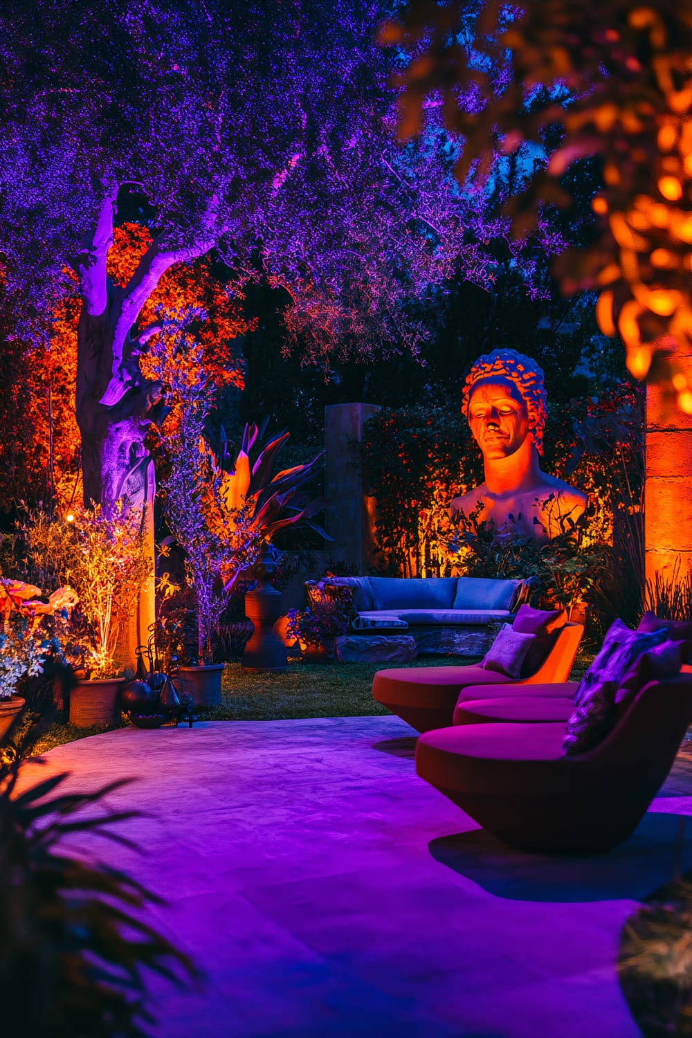 An eclectic outdoor garden decorated for Halloween with vibrant orange and purple lighting. The garden features artistic sculptures, including a large bust of a man, and bold floral arrangements with dark foliage. Stylish seating areas, including red round chairs and a stone bench with cushions, are spread across the space. The dynamic lighting creates a lively and sophisticated nighttime setting.