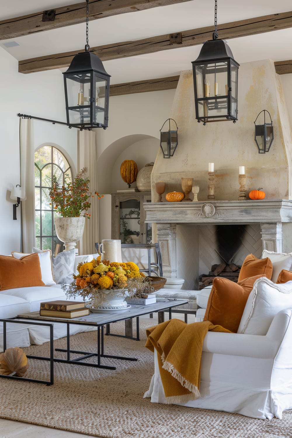 A beautifully decorated living room with a rustic feel. The room features high wooden beams, two large black lantern-style chandeliers, and an impressive, weathered stone fireplace adorned with pumpkins and various wooden candlesticks and decorative objects. The seating area includes white sofas and armchairs with orange and mustard-colored cushions and blankets, centered around a light grey coffee table displaying a vibrant floral arrangement and books. A large, woven jute rug covers the floor. The background showcases arched windows with flowing cream curtains, allowing natural light to flood the room. A tall, classic planter with autumnal foliage adds a touch of nature.