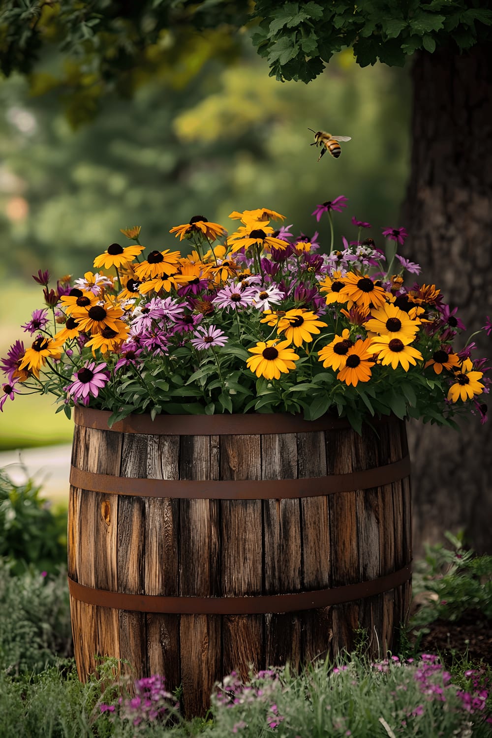 A repurposed wooden barrel planter overflows with a vibrant mix of Black-Eyed Susans, Purple Coneflower, Shasta Daisies, and Scarlet Bee Balm, caught in the golden glow of summer afternoon light. A single honeybee, frozen in mid-flight, hovers near the flowers. In the background, soft light filters through the dappled foliage of nearby trees.