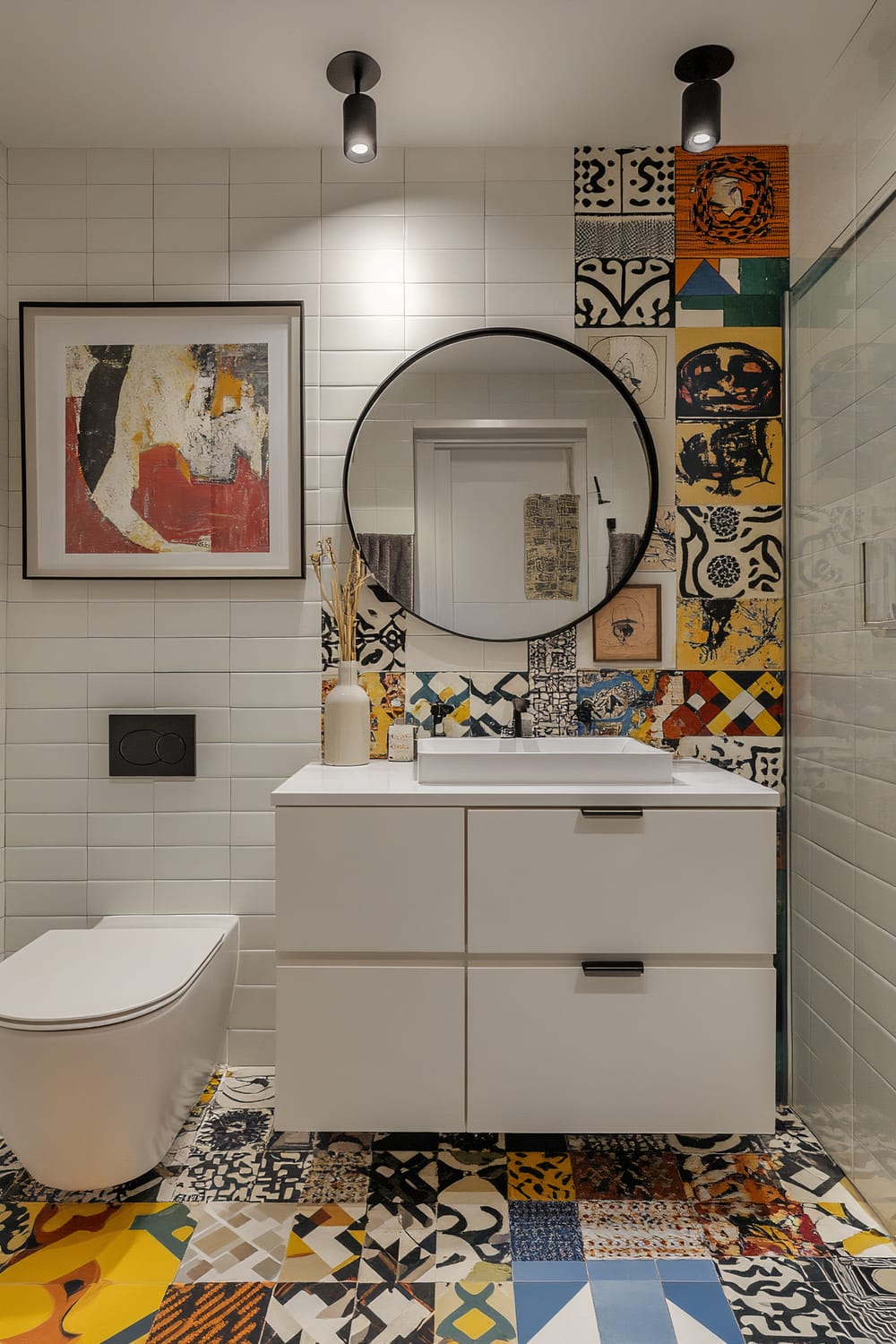 This bathroom features a striking combination of minimalist and eclectic design elements. The walls are tiled with plain white rectangular tiles, while one section showcases a vibrant patchwork of multicolored, patterned tiles extending from the floor. A suspended, modern white vanity with a square sink sits below a large circular mirror. Above the toilet hangs an abstract framed painting with red, black, and white hues. The space is softly lit by black ceiling-mounted lights.