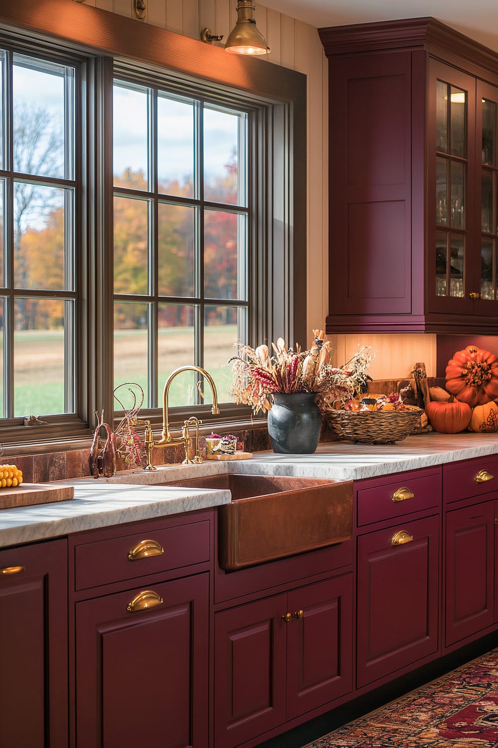 A kitchen scene features deep maroon cabinetry with brass handles and cream-colored countertops. A copper farmhouse sink is installed under a large set of windows, which display a view of autumn foliage outside. The countertop is adorned with decorative items like a black vase filled with dried plants, a basket containing fall-themed ornaments, and a small arrangement of pumpkins. Overhead lighting casts a warm glow, highlighting the kitchen's rustic elegance. A colorful, patterned rug lies on the floor, adding to the cozy ambience.