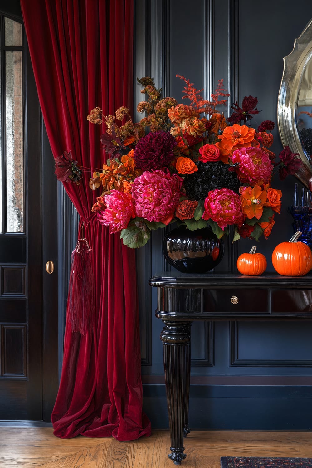 A luxurious interior scene with dark, glossy walls and a polished wooden floor. A black side table with fluted legs holds a large floral arrangement featuring vibrant red, pink, orange, and burgundy flowers in a black vase. Two small orange pumpkins are placed on the side table, adding a touch of seasonal decor. A rich, red velvet curtain frames one side of the scene, creating a dramatic backdrop.