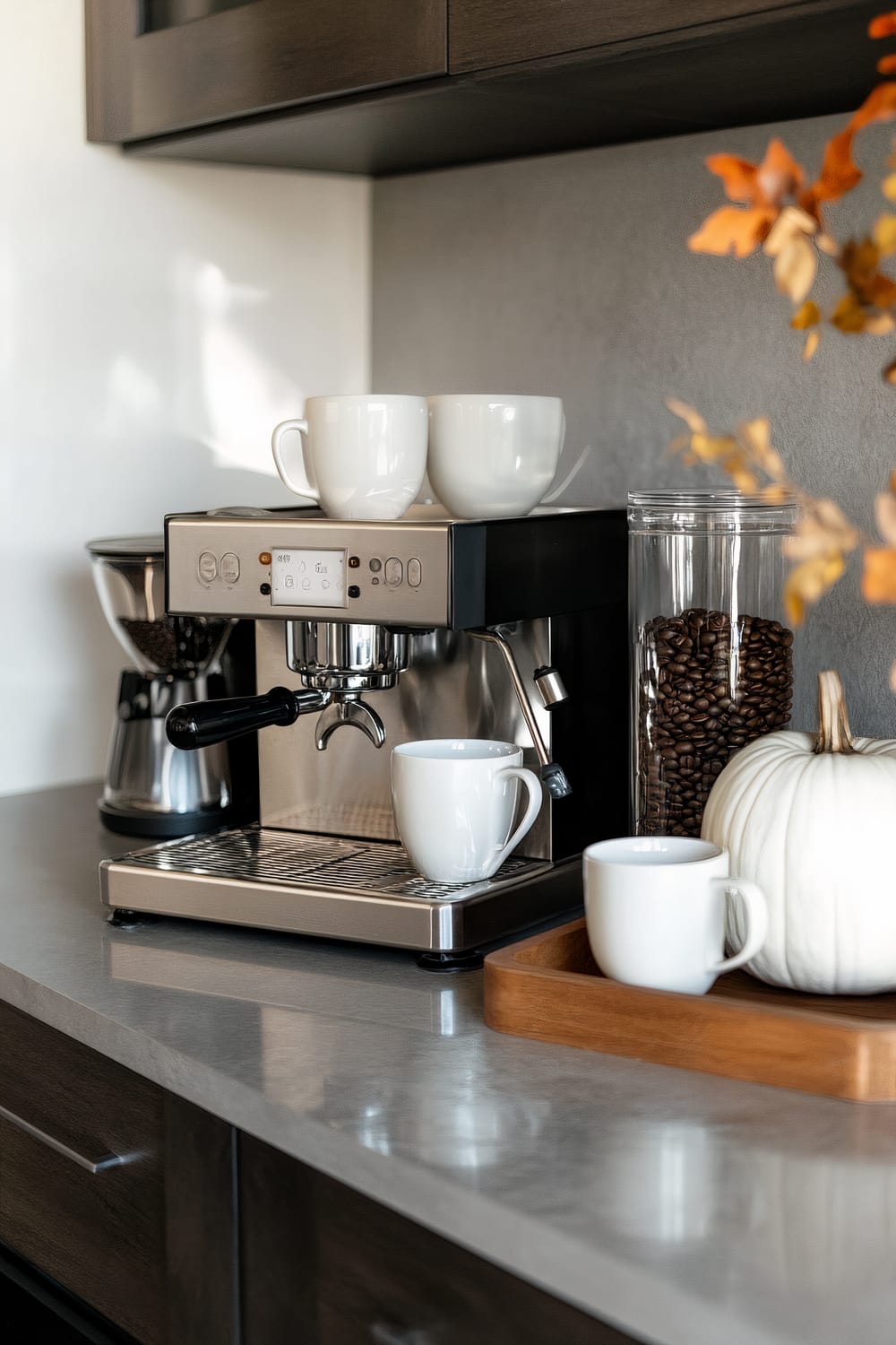 A modern kitchen counter displays a sleek espresso machine with white ceramic cups placed on top and under the coffee spout. Adjacent to it, there is a coffee grinder and a large glass jar filled with coffee beans. A small wooden tray holds a white ceramic cup and a white ornamental pumpkin. Fall leaves in the background add a touch of seasonal decor.