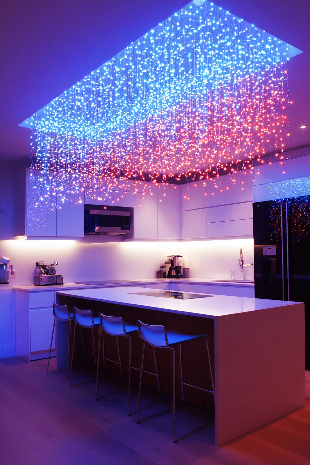 A spacious kitchen with a ceiling covered by cascading multi-colored fairy lights, forming a starry pattern. Below, a sleek kitchen island with mirrored surfaces reflecting the vibrant lights. The kitchen has white cabinetry, modern appliances, and stools on one side of the island.