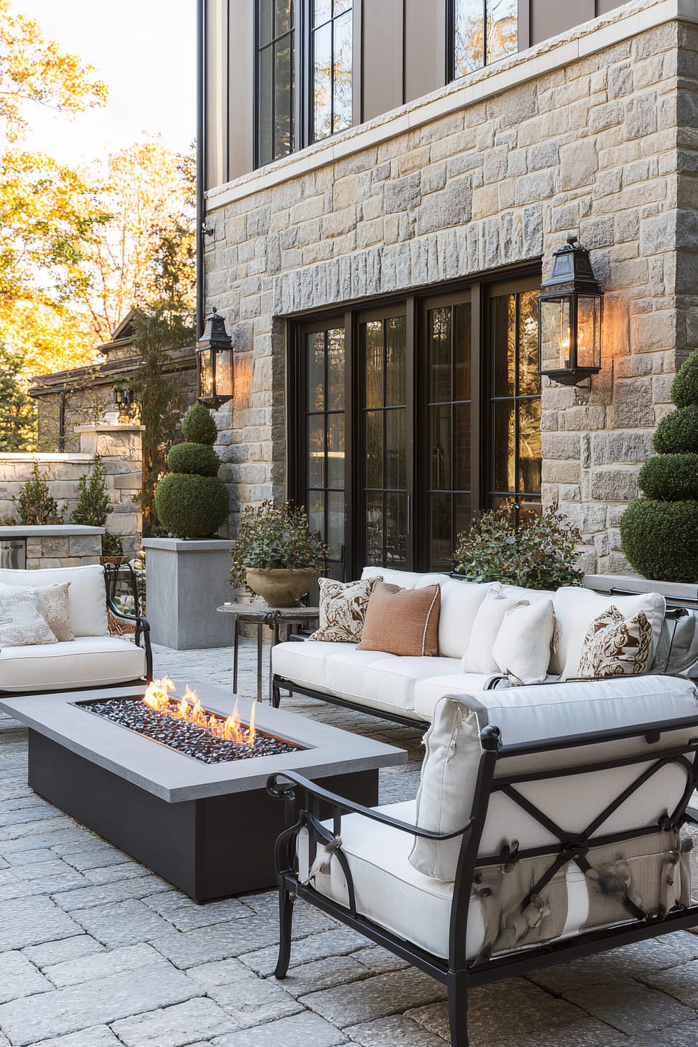 An outdoor patio featuring elegant furniture with white cushions, a modern rectangular fire pit, and well-maintained topiary plants. The patio is bordered by a stone facade with large windows and classic outdoor wall lanterns. Potted plants add a touch of greenery.