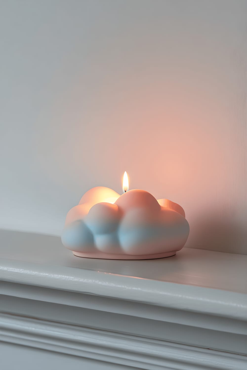 A pastel-colored cloud-shaped candle holder on a white minimalist mantelpiece. The candle holder transitions from pink to blue and holds a gently glowing candle, creating a soothing ambiance.