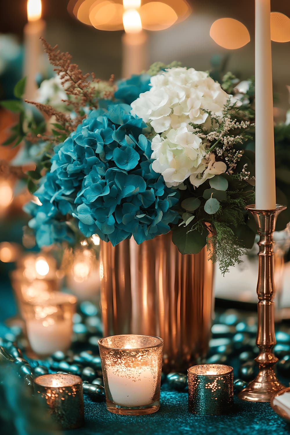 A dramatic table centerpiece composed of a large copper vase holding vibrant teal hydrangeas and trailing ivy. The vase is surrounded by smaller copper candlesticks with white taper candles and teal glass beads scattered around. These elements are beautifully arranged on a rich emerald green table runner with a subtle metallic sheen, basking under warm, angled light.