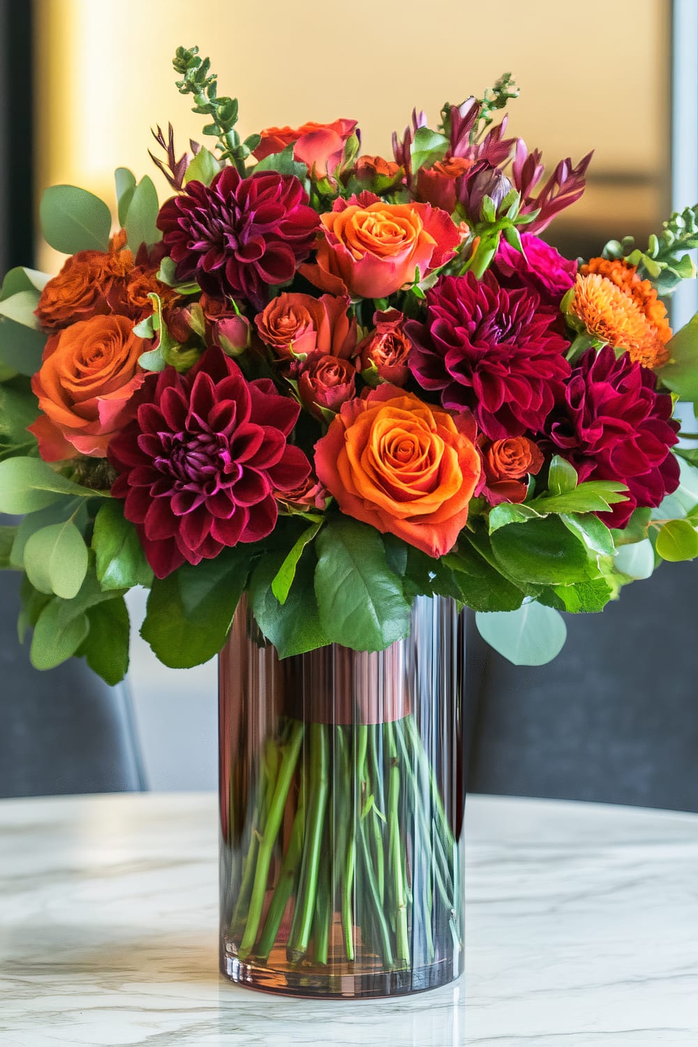 A vibrant bouquet of flowers featuring deep red dahlias, orange roses, and other assorted blooms in a tall, clear glass vase. The arrangement is displayed on a marble surface with a blurred background, emphasizing the floral centerpiece.