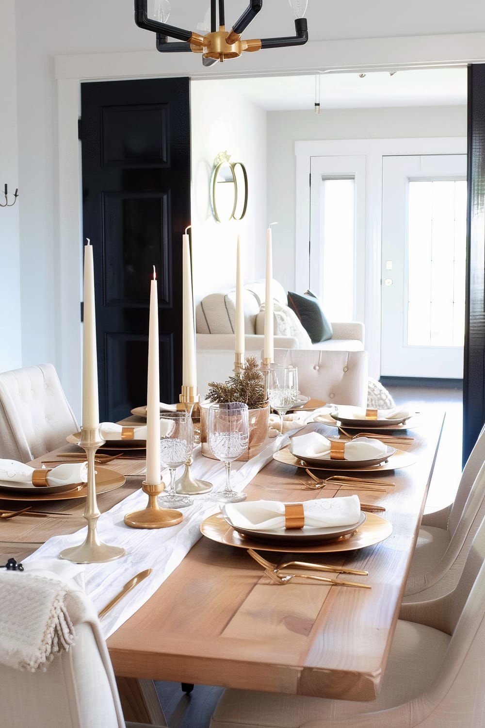 This image features a sophisticated dining table setup for a formal occasion. The wooden table is adorned with a white table runner, and each place setting includes a gold charger plate, white napkin with a gold napkin ring, and gold utensils. Crystal glasses are set for each guest, and tall white candles in gold candlesticks are arranged down the center of the table. The space is illuminated by a modern chandelier with black and gold finishes. The background shows a seating area with a cream sofa and stylish accents, including a black door and a decorative sconce on the wall.