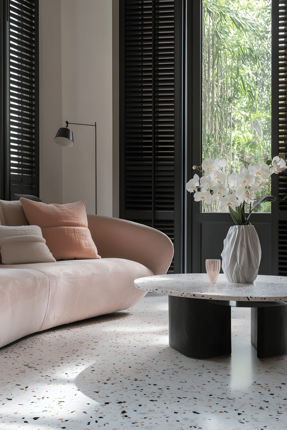 A spacious Milanese living room with asymmetrical design elements. The room features terrazzo flooring in shades of white, grey, and subtle pink with a large abstract terrazzo coffee table in the center. One wall is adorned with a soft blush pink asymmetrical sofa, accompanied by a black lacquered side table. A minimalist floor lamp with a matte black base and white shade stands near the sofa, and a modern sculptural vase with white orchids decorates the room. The room is well-lit by natural light from the large floor-to-ceiling windows with dark grey shutters, creating intricate shadows on the terrazzo floor.