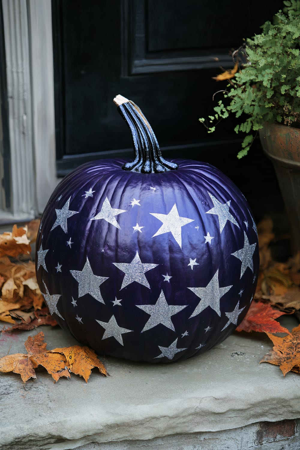 A dark purple pumpkin with a glossy finish sits on a stone surface against a dark background. The pumpkin is adorned with silver glitter stars of various sizes, giving it a celestial appearance. The surroundings include scattered brown and orange autumn leaves and a potted green plant partially visible to the right.