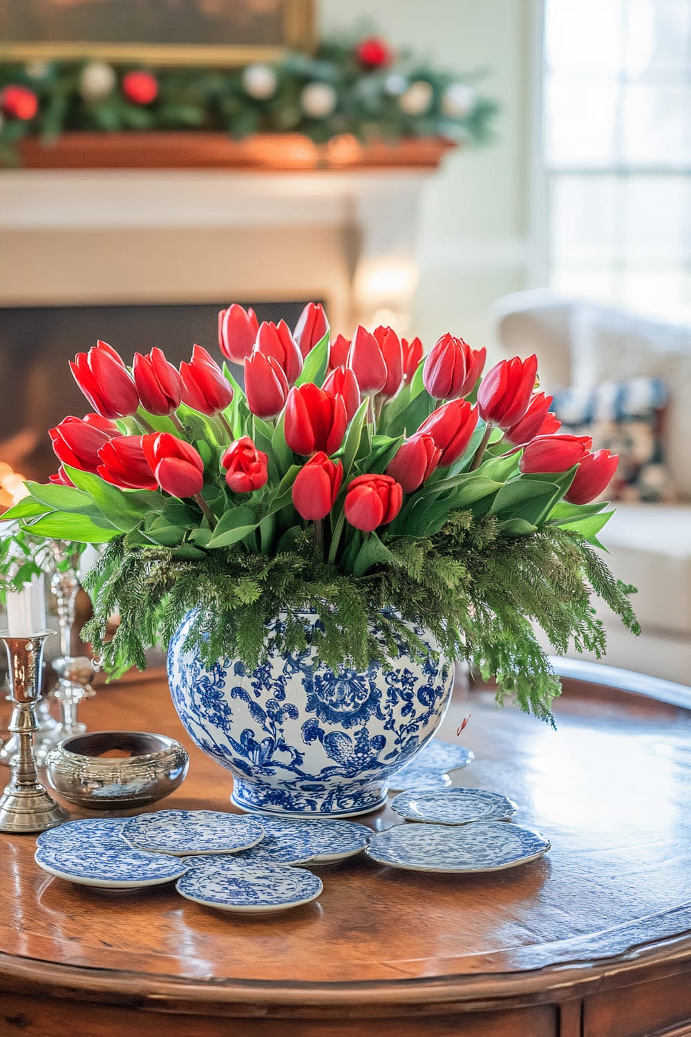 An ornate vase filled with red tulips sits on a wooden coffee table. The vase is blue and white with intricate floral patterns. Surrounding it on the table are multiple blue and white decorative plates. In the background, there is a fireplace mantel adorned with green garland and red and white ornaments, and a partially visible armchair next to a window letting in daylight.