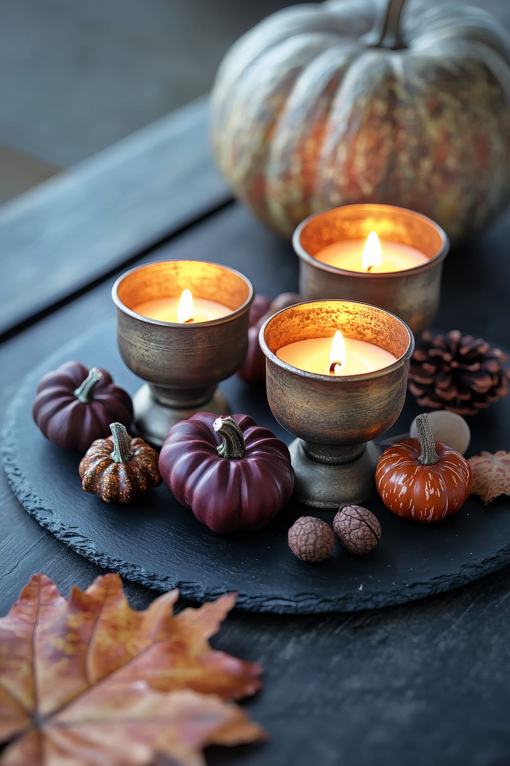 Three lit candles in elegant chartreuse holders are arranged on a black slate plate. Surrounding the candles are decorative burgundy elements including small pumpkins and acorns. A large pumpkin in the background and autumn leaves in the foreground contribute to the fall-themed decor.