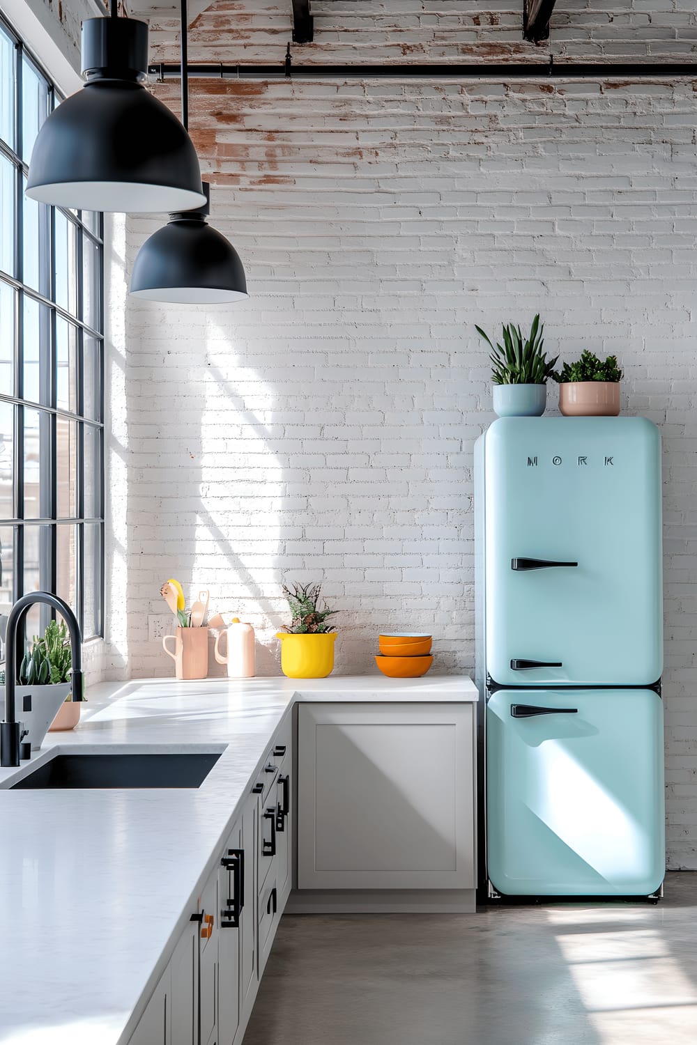 An open-concept loft kitchen featuring white exposed brick walls, light gray concrete flooring, and large industrial-style windows that fill the room with natural light. A white marble countertop is located in the center of the space, adorned with black matte fixtures and a collection of colorful ceramic mixing bowls. Above the countertop hang pendant lights with matte black metal finishes. A retro-styled refrigerator with pastel accents is tucked against the wall, and a potted succulent sits in a modern geometric planter on the countertop.