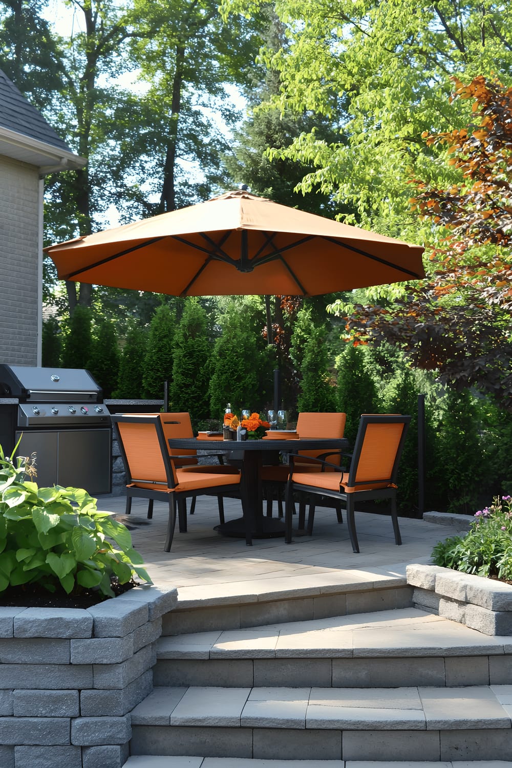 A stylish backyard grill area with a semi-circular stone patio. The space is adorned with a black dining table surrounded by four vibrant orange chairs under a large brown umbrella. A functional built-in stone grill station with countertop space is present, while meticulously trimmed shrubs and a blooming tree provide a serene, landscaped backdrop. The area appears to be harmoniously integrated with the house's outdoor design.