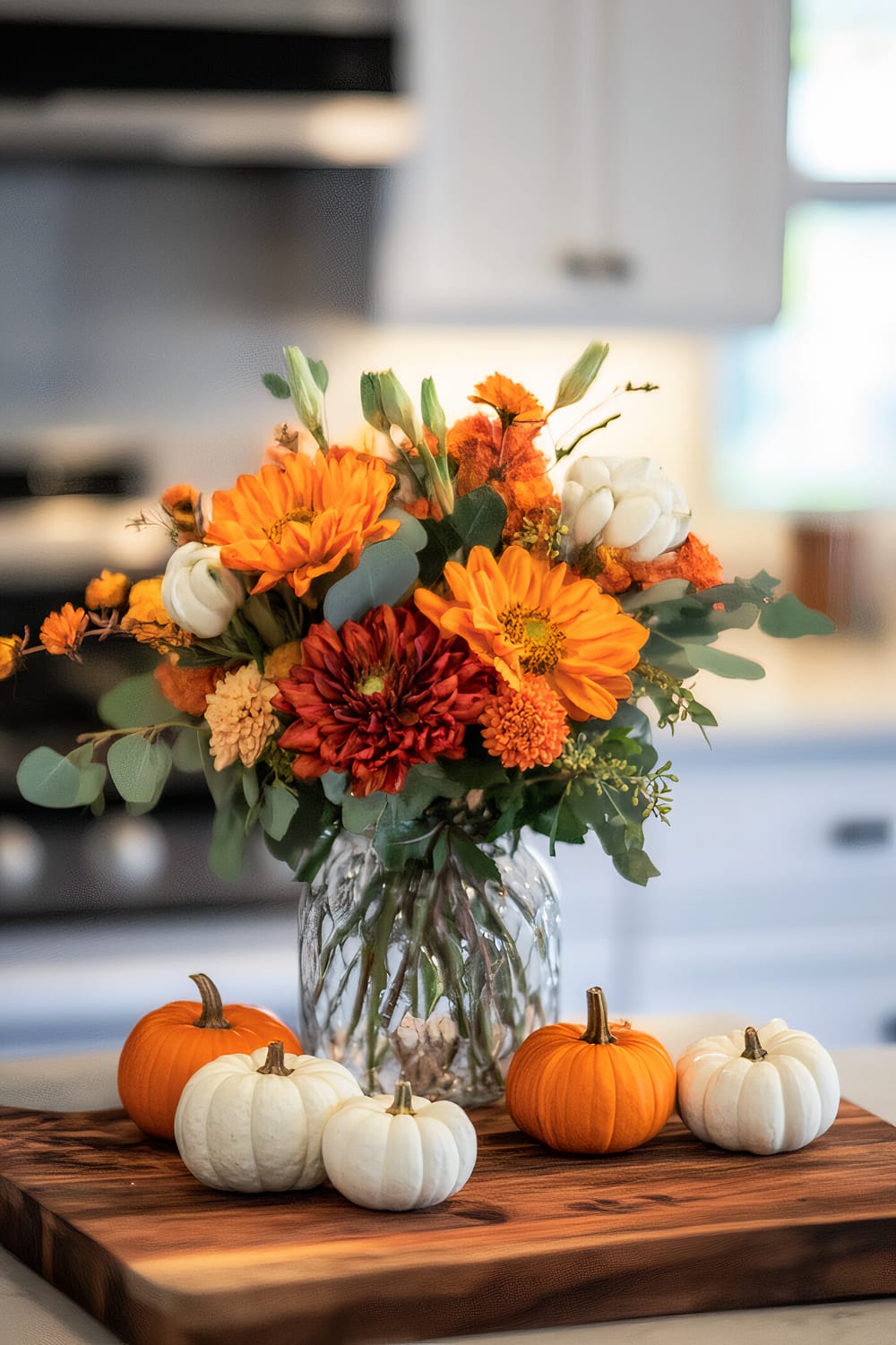 A bright and vibrant floral arrangement in a clear, textured glass vase, placed centrally on a wooden cutting board in a kitchen. Surrounding the vase on the board are small decorative pumpkins in white and orange, adding to the autumnal theme. The background features modern white cabinetry and a stovetop, softly blurred to keep the focus on the centerpiece.