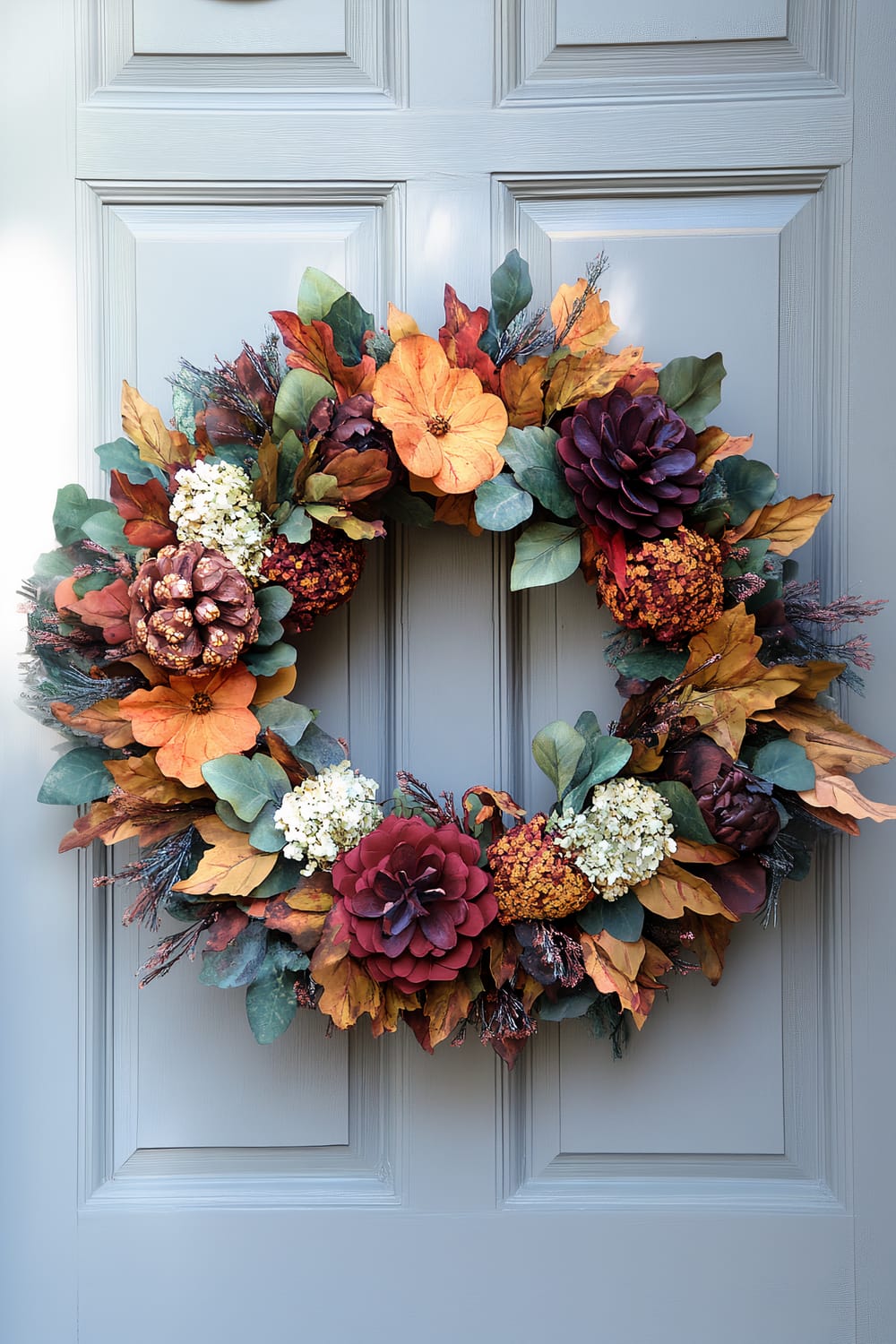 A festive and lush autumn wreath adorns a soft grey-painted front door. The wreath features an array of fall foliage in vibrant hues of orange, red, yellow, and green. It includes elements such as pinecones, artificial flowers, and various types of leaves, all meticulously arranged to showcase the essence of fall. The grey door contrasts beautifully with the rich autumn colors of the wreath, enhancing its visual appeal.