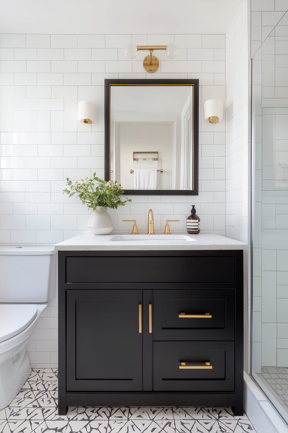 This image depicts a modern bathroom featuring a sophisticated black vanity with gold hardware and fixtures. The vanity is topped with a sleek white countertop, adorned with a simple yet elegant white vase containing green foliage and a brown bottle beside the sink. Above the vanity hangs a rectangular mirror with a black frame and gold inner trim, complementing the vanity. The wall behind the vanity showcases white subway tiles arranged in a clean, grid pattern, offering a crisp and fresh backdrop. On either side of the mirror are sleek wall-mounted sconces with white cylindrical shades and gold bases, adding a touch of luxe to the space. The floor is covered in geometric patterned tiles in black and white, adding visual interest. To the left of the vanity, a white toilet is visible, and on the right, there is a corner of a glass shower door, suggesting a well-integrated bathroom design. Over the mirror, a gold light fixture with two bulbous glass shades is mounted, completing the bathroom's modern and elegant aesthetic.