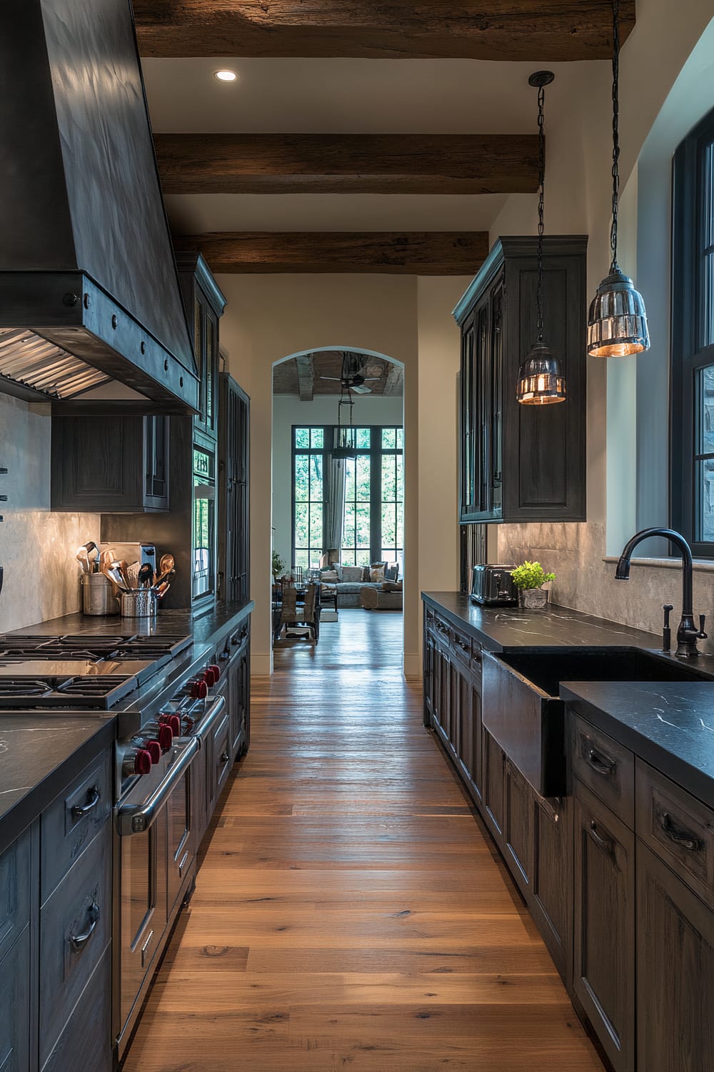 A moody farmhouse kitchen viewed from a hallway archway. The kitchen features deep charcoal cabinetry with dark walnut countertops creating a refined atmosphere. There is a stainless steel range and an espresso machine on the black marble countertop. Dramatic overhead lighting with dark metal fixtures and exposed wooden beams add a rustic charm. The textured stone backsplash enhances the space's sophistication.