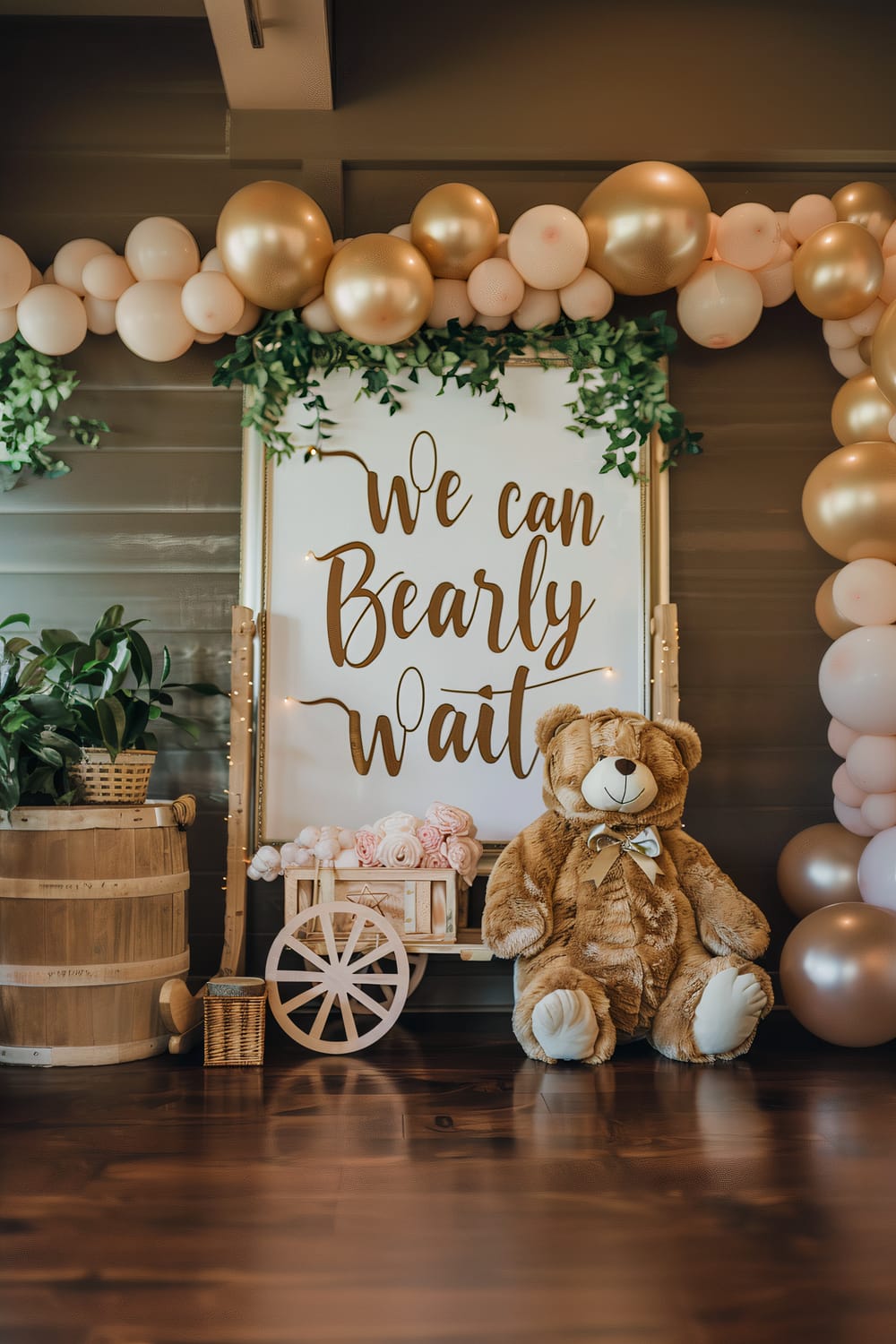 A beautifully decorated nursery corner features a prominent sign with the words "We can Bearly Wait" in elegant script. Surrounding the sign are clusters of balloons in white, gold, and blush tones, interspersed with green foliage. A large, plush teddy bear with a bow sits beside a small wooden cart filled with rolled baby blankets. Next to the cart is a rustic wooden barrel with a green plant in a wicker basket on top. The scene is set against a wall with dark horizontal panels and polished wooden floor.