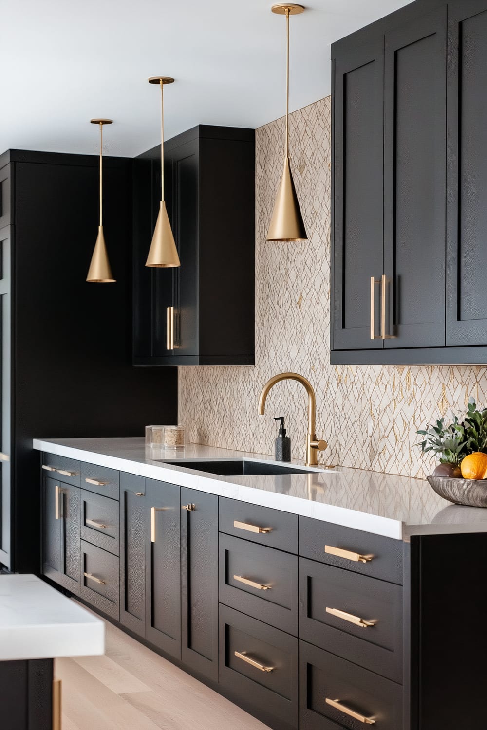 A modern kitchen with matte black cabinets, adorned with gold handles, and a white marble countertop. The backsplash features a textured, geometric pattern in beige and gold. Three gold, cone-shaped pendant lights hang above the counter. A gold faucet is installed over the sink, and a bowl of fruits with green foliage is placed on the countertop.