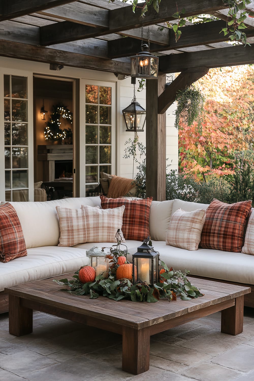 An outdoor seating area under a wooden pergola with hanging lanterns. There is a large L-shaped white cushioned sofa with plaid throw pillows in autumn colors. A low wooden coffee table is decorated with pumpkins, greenery, and lanterns. The background shows a house with large windows and seasonal foliage.