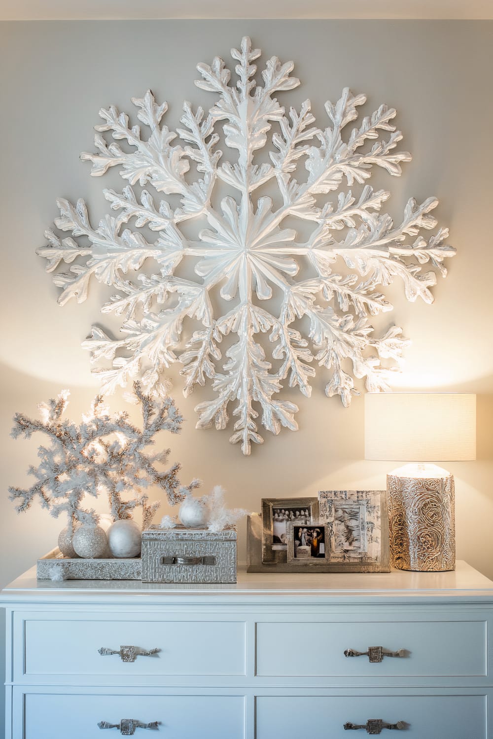 A large white porcelain snowflake mirror is mounted on the wall above a white dresser in a bedroom. On top of the dresser, there are various decorative items including a small white tree adorned with white ornaments, a set of picture frames, and a stylish lamp with a patterned base. The dresser features modern metal handles, and the room is lit with both natural and ambient lighting, creating a festive and elegant ambiance.