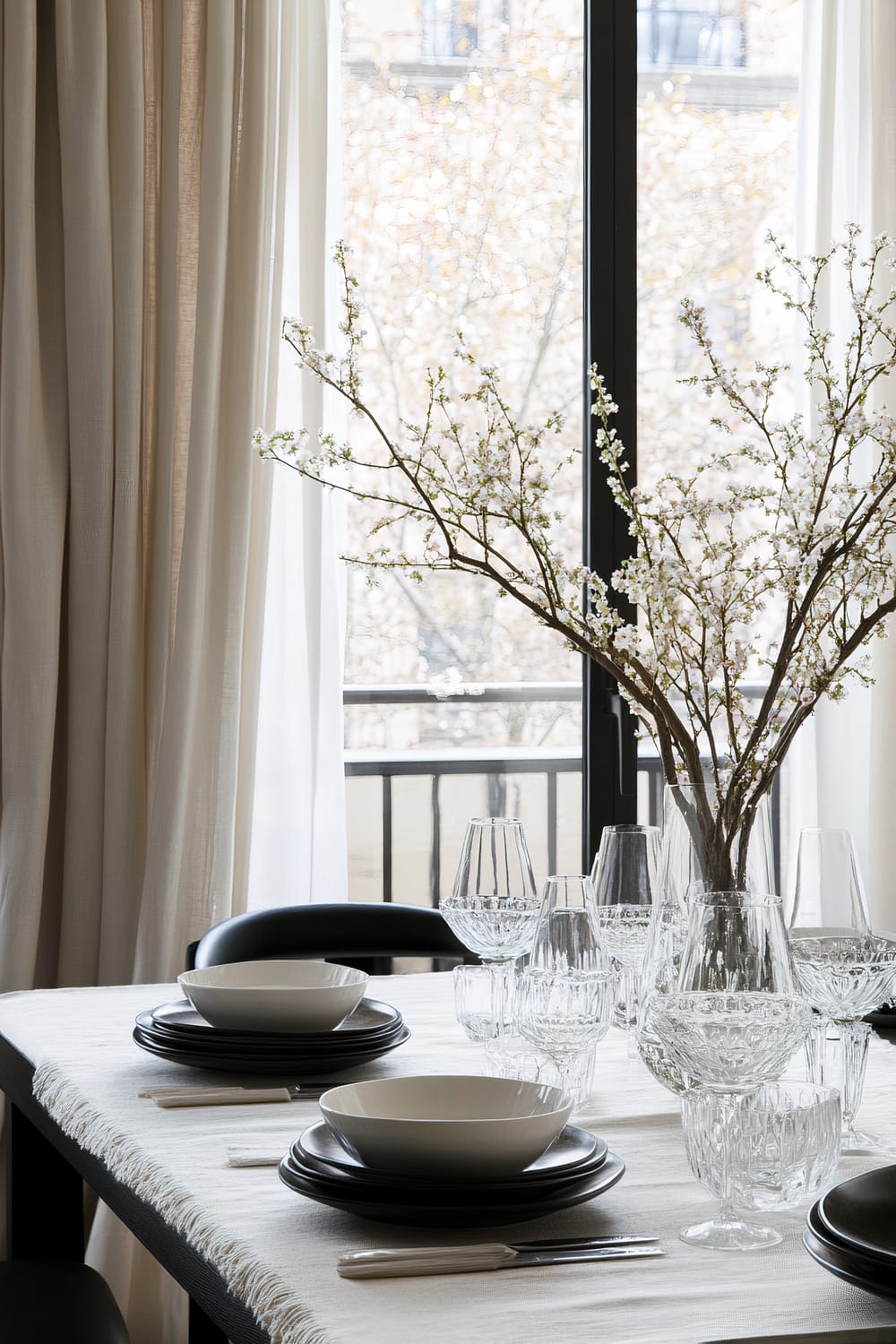 A modern dining table is adorned with black and white place settings, featuring black plates and white bowls, each with white-handled cutlery alongside. In the center of the table, there's a tall vase filled with branches of delicate white blossoms. Various crystal glassware is meticulously arranged, adding a touch of elegance. Behind, large windows with beige curtains provide a view of a softly blurred, tree-lined urban street, allowing natural light to flood the room.