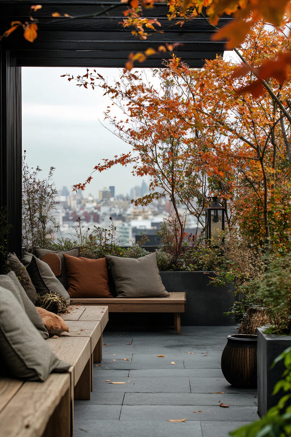 A rooftop terrace featuring wooden benches with earth-toned cushions under a pergola with a city skyline visible in the distance. The scene is adorned with autumnal trees, their orange leaves providing a vibrant contrast to the gray cityscape and potted plants that enhance the serene ambiance.