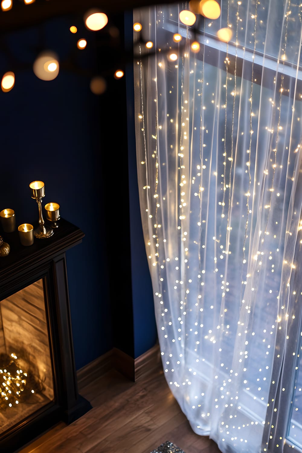 An overhead view of a festively decorated living room. A deep navy wall provides the backdrop for warm, starry white fairy lights draped around large window frames. In the center of the room, a mantelpiece houses three gold candle holders and specks of gold confetti scattered on its surface.