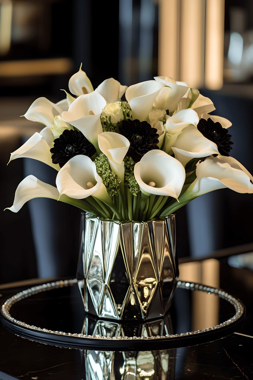 A lavish Art Deco-inspired centerpiece placed on a kitchen table, featuring a balanced arrangement of white calla lilies and black dahlias in a geometric mirrored vase. There are small shiny gold accents and crystal beads enveloping the base of the flowers. This whole arrangement is placed on a sleek black marble tray carried by a navy velvet fabric, under focused, warm lights that bring an extra gleam to the metallic and mirrored surfaces of the decor.