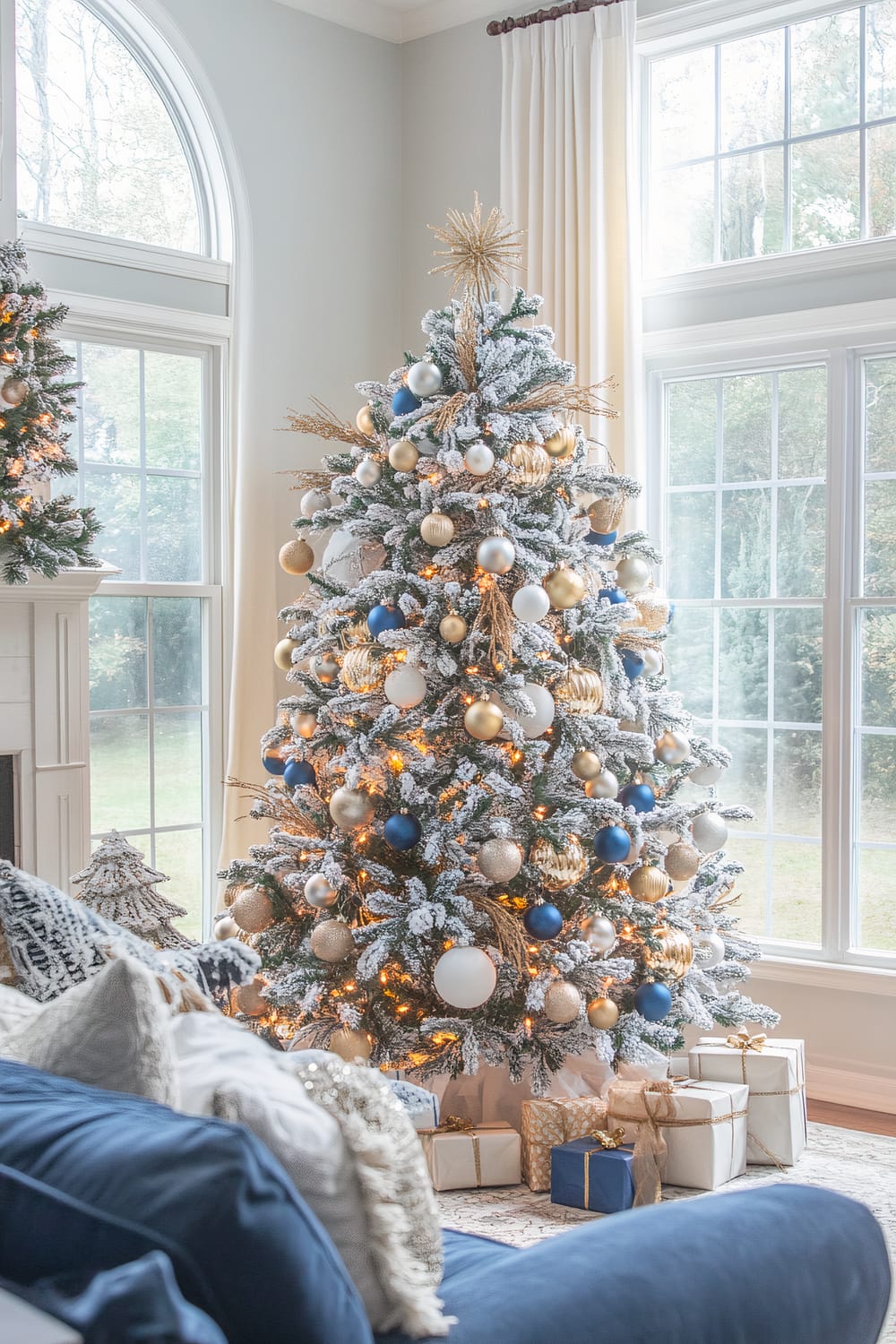 A living room decorated with a Christmas tree that reaches up to the ceiling, adorned with a mix of gold, white, blue, and silver ornaments, and warm lights. The tree has a snowy frost effect and a starburst tree topper. Below it are neatly arranged wrapped gifts in gold, white, and blue wrapping papers. The room features large windows with white curtains, lending ample natural light, and a blue sofa with cream and blue pillows and blankets. A smaller, similarly decorated green tree is partially visible by the mantel.