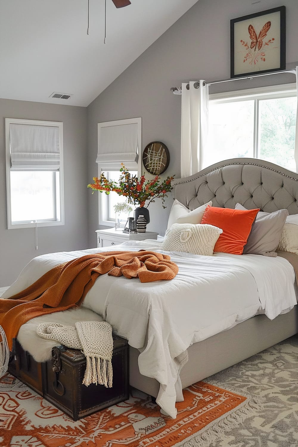 A stylish bedroom featuring a gray upholstered bed with tufted headboard adorned with white, gray, and orange pillows. An orange blanket is draped casually over the bed, which has white bed linens. A vintage chest at the foot of the bed holds a knitted cream throw. Beside the bed, a white nightstand with a dark vase containing a bouquet of colorful flowers is situated. The room has gray walls, white-framed windows with Roman shades, and a tall ceiling with a ceiling fan. Artwork including a butterfly print and abstract piece decorates the walls.