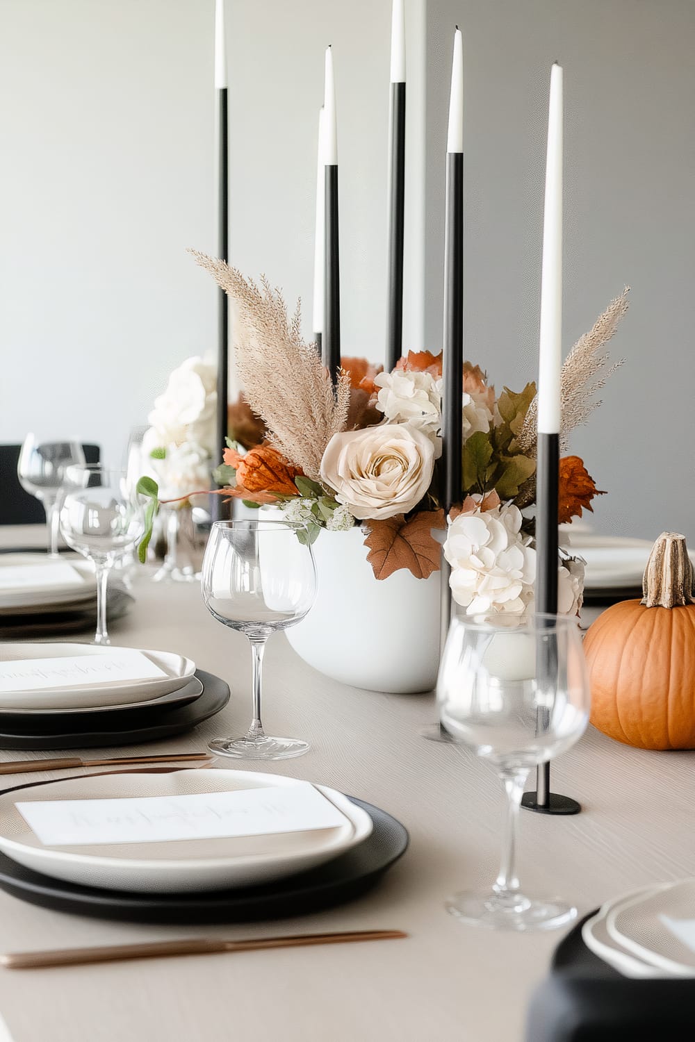 An elegantly set dining table features black and white tall candles, placed in black holders, that are complemented by an arrangement of white, beige, and orange flowers and foliage, including a small pumpkin. The table is set with black and white plates, clear wine glasses, and sleek cutlery.