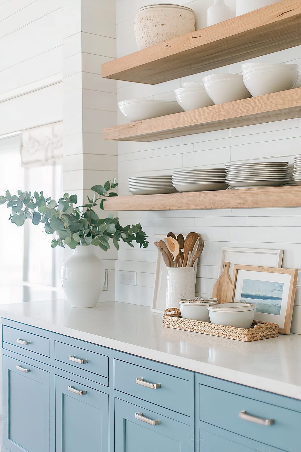 A modern kitchen counter with a clean and minimalistic design. The cabinetry is painted in a soft blue hue with sleek silver handles. On the white quartz countertop, there's a large white vase filled with green eucalyptus branches. Two floating wooden shelves mounted on the white tiled backsplash hold neatly arranged white bowls and plates. Below the shelves, there are various kitchen utensils in a white ceramic holder, a small framed picture, a wooden cutting board, and ceramic dishes on a woven tray.